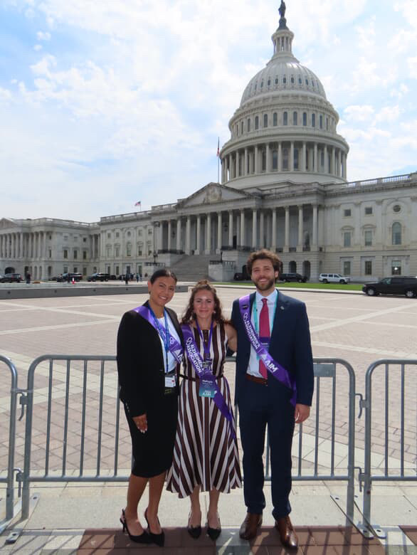 Three students pose together in front of U.S. Capitol Building. Over their clothes, each is wearing a purple sash for Alzheimer's advoacy
