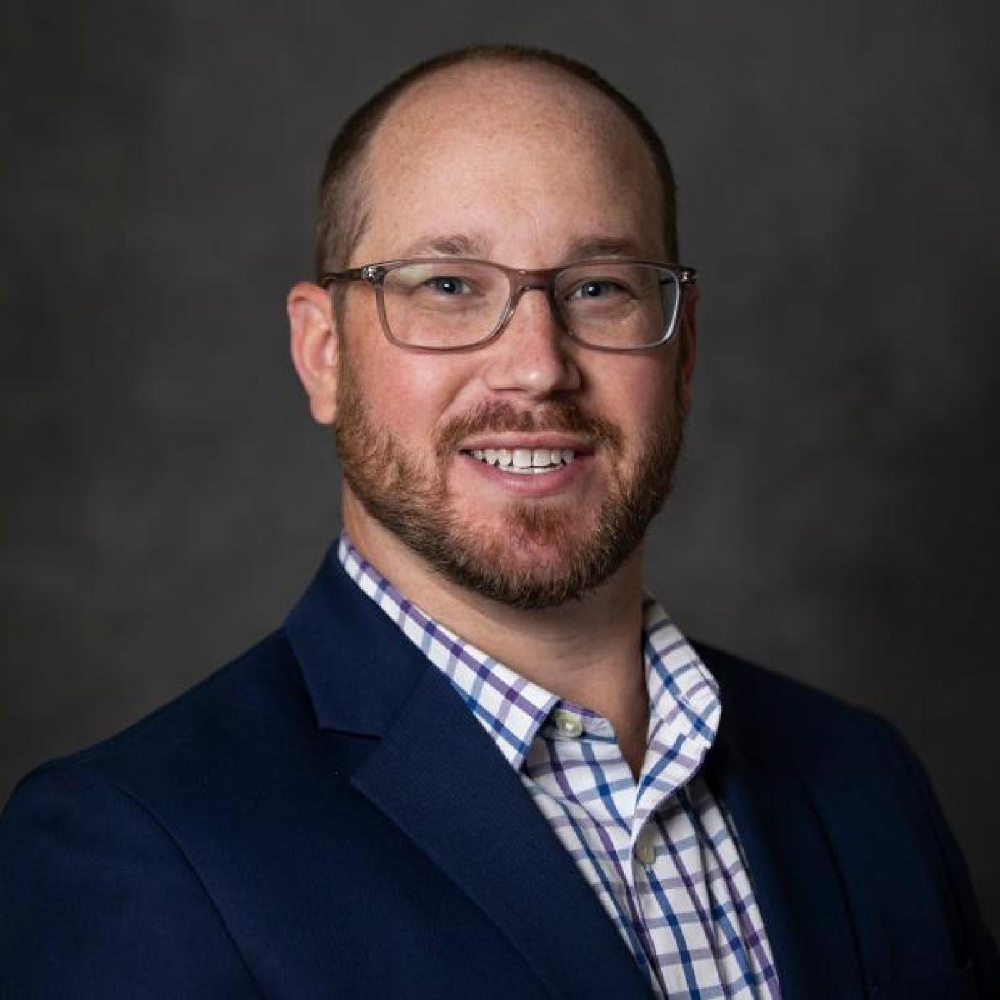 Kirby Mayer smiles in his headshot. He wears a blue jacket and checkered button down.