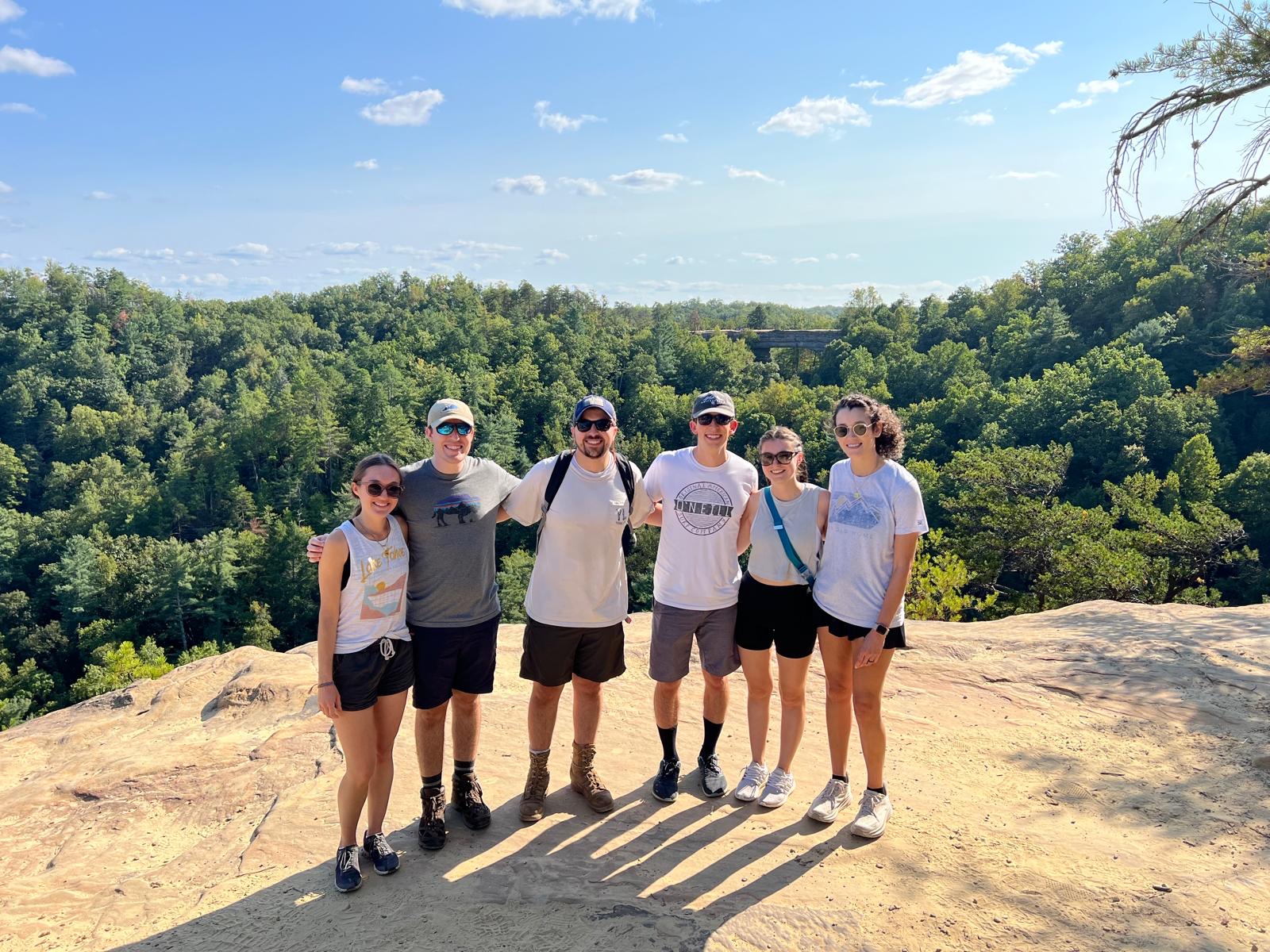 Group of six residents smiling on a hike, standing on a rocky ledge with lush green forest and blue sky in the background, conveying adventure and camaraderie.