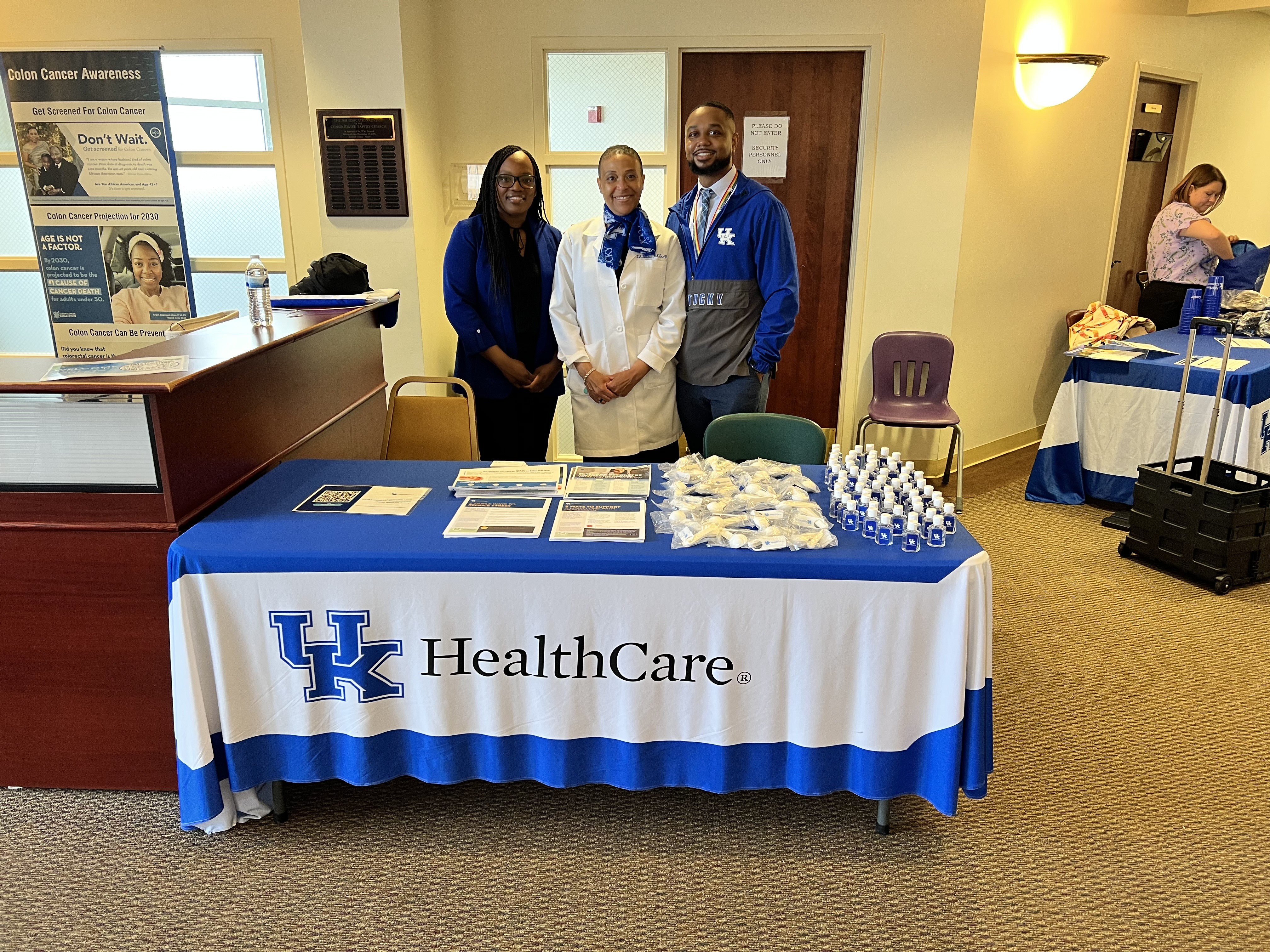 Three individuals stand at a table titled "UK HealthCare" and smile at the camera