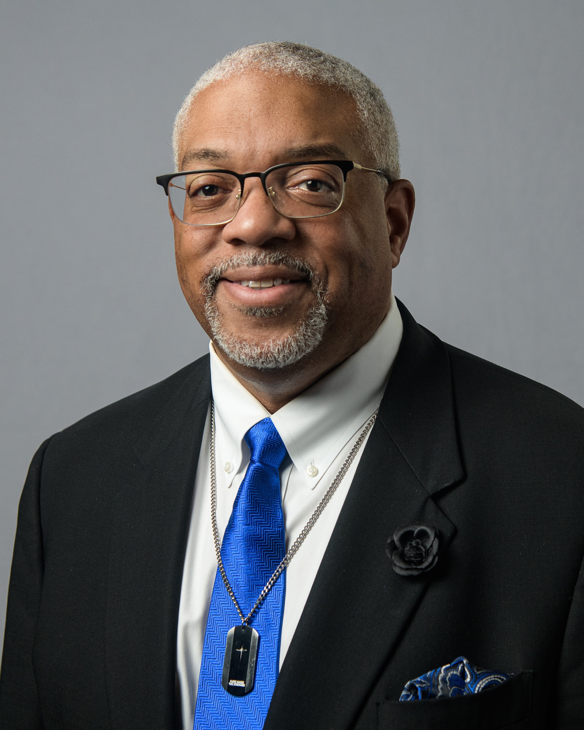 A headshot of a man in a black suit and blue tie with a silver dog tag around his neck