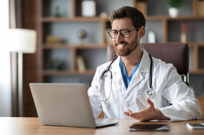 A man with dark hair, a beard, and glasses, wearing a white coat while holding a web call.