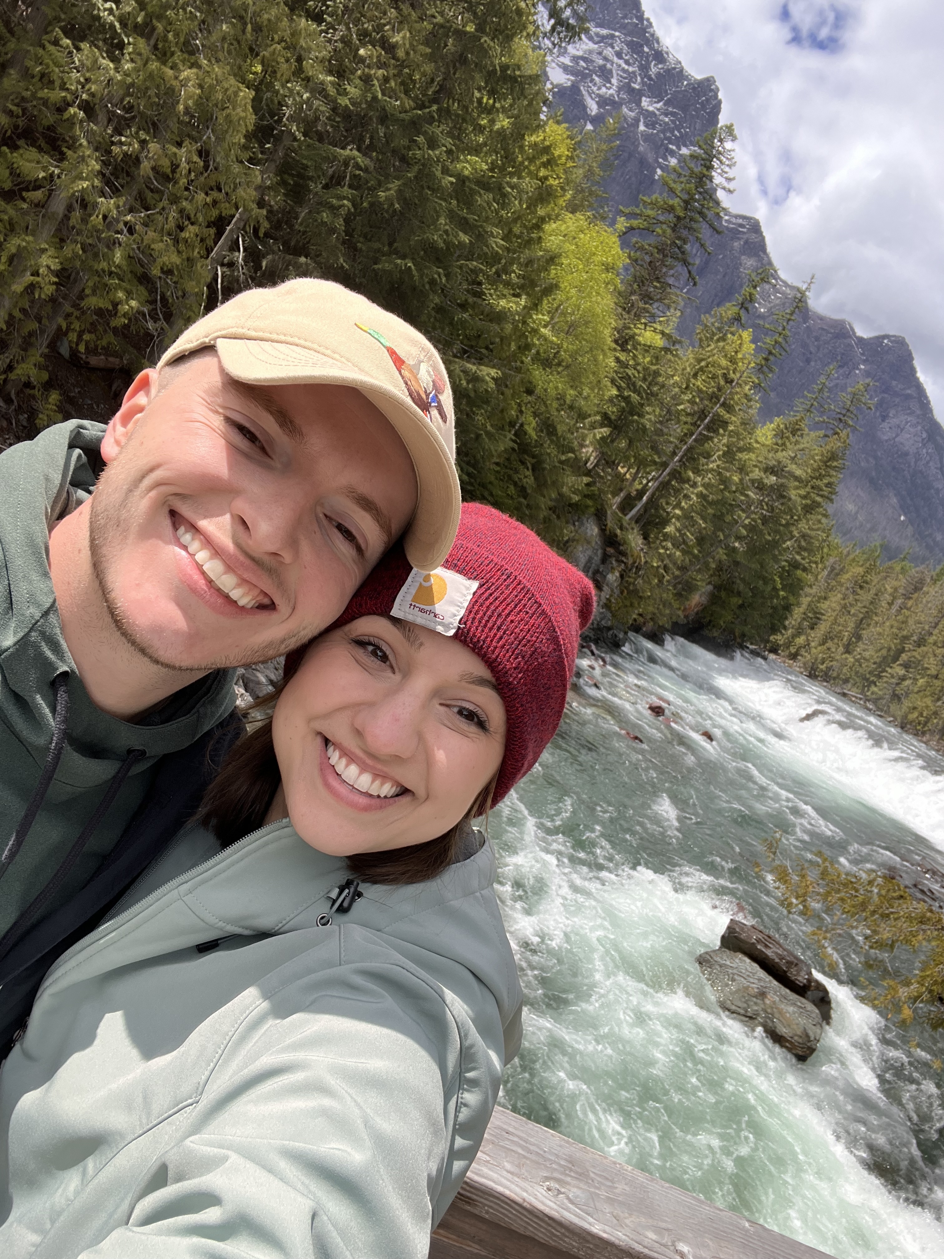 Anna cox and her husband taking a selfie on a wooden bridge over a rushing river, surrounded by lush greenery and mountainous landscape under a partly cloudy sky.