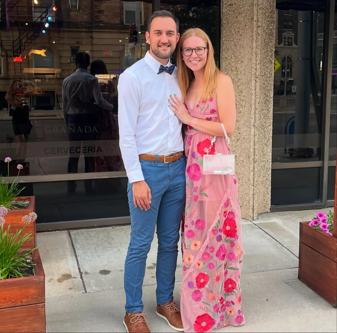 Thomas Calderaro and his significant other standing in front of a restaurant. She wears a floral dress and glasses, and he is in a white shirt and bow tie. Flower boxes flank them.