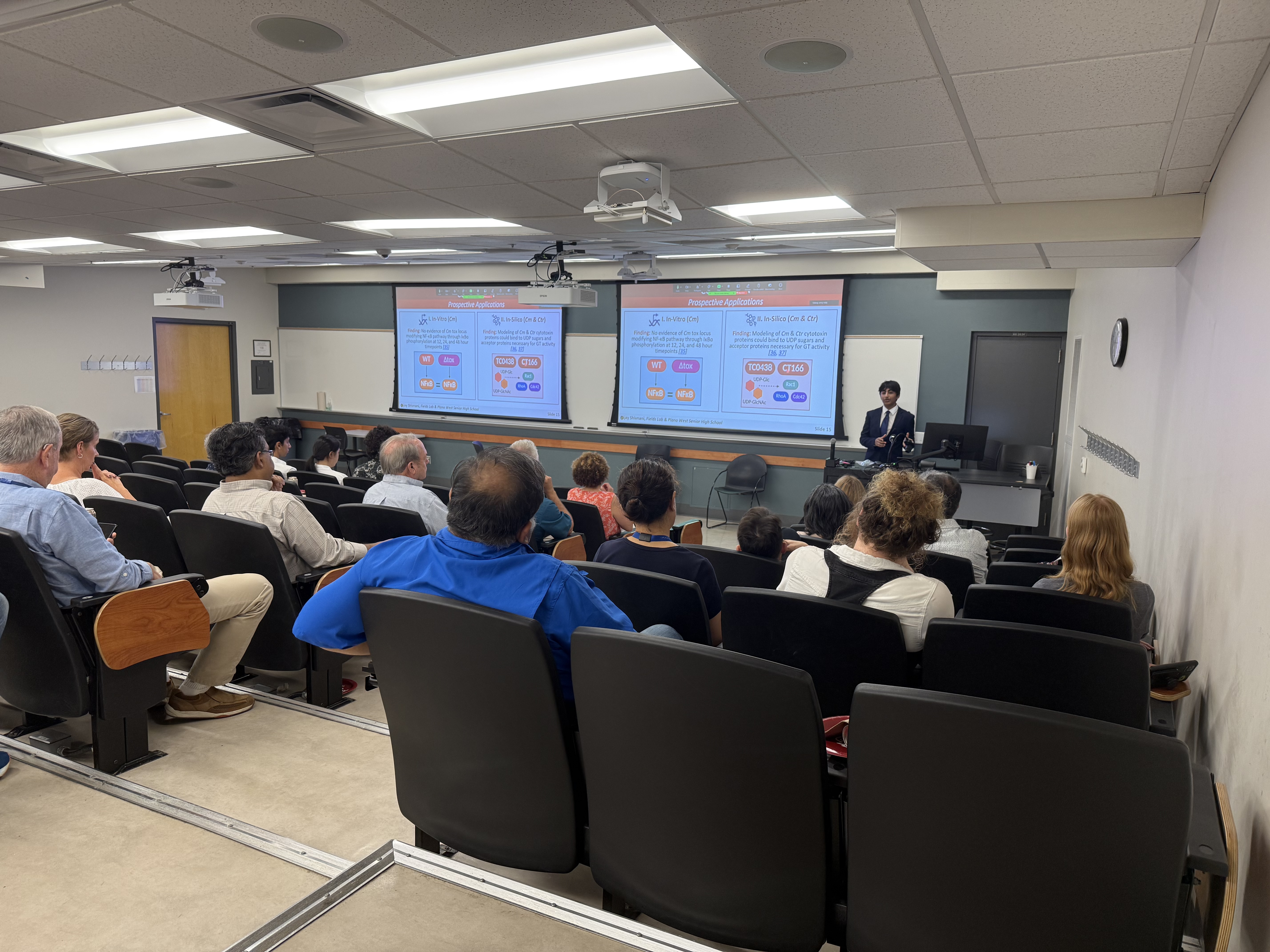 A male presenting high school student gives presentation in front of seated audience. Screens with projected data appear behind him.