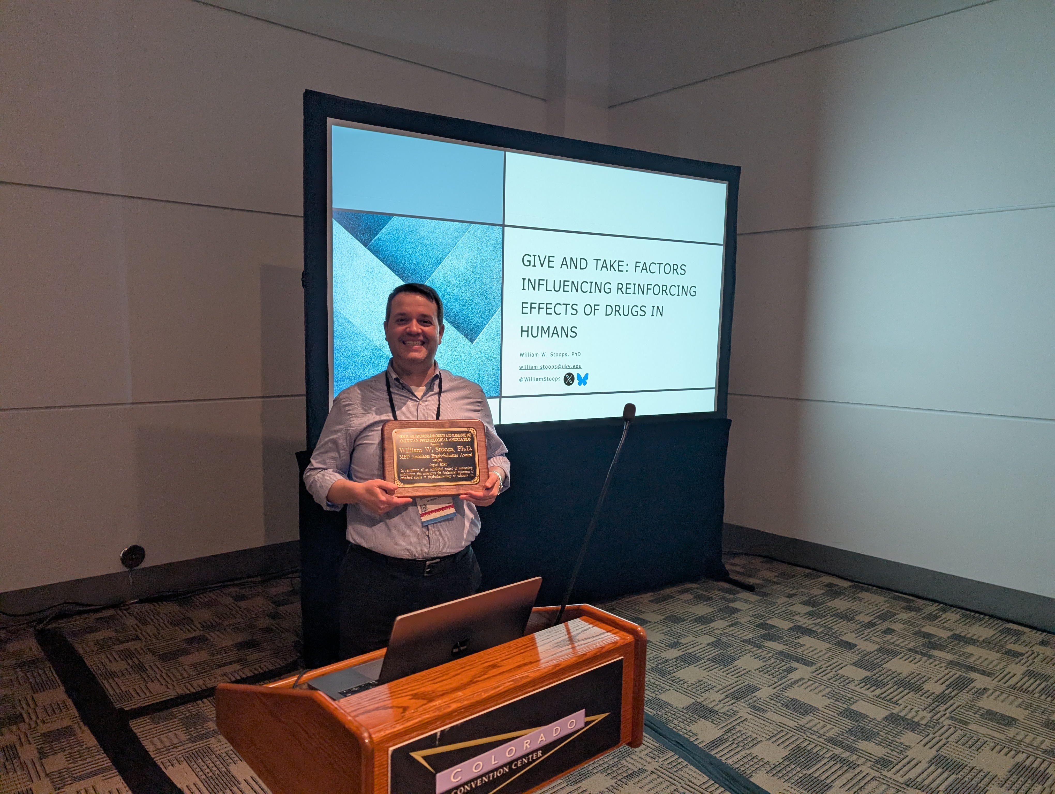 Dr. Stoops poses in front of a projected screen, holding his plaque