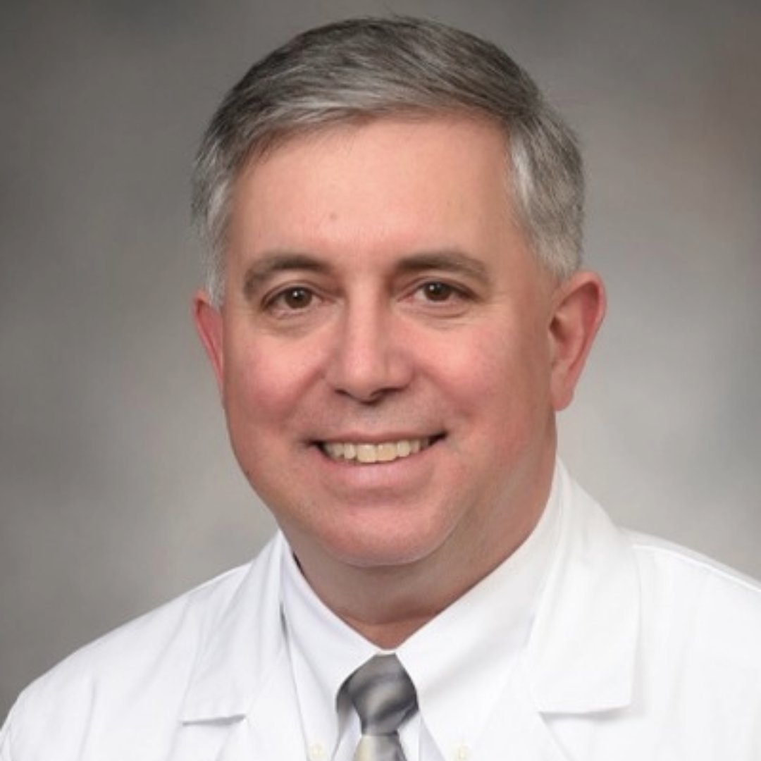 A man in a white coat and gray tie smiles for a headshot