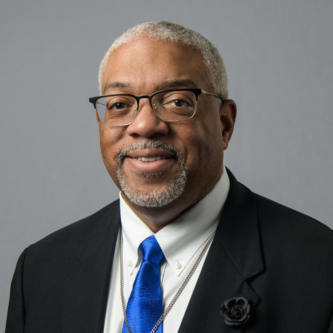 A man in a black suit and blue tie smiles for a headshot