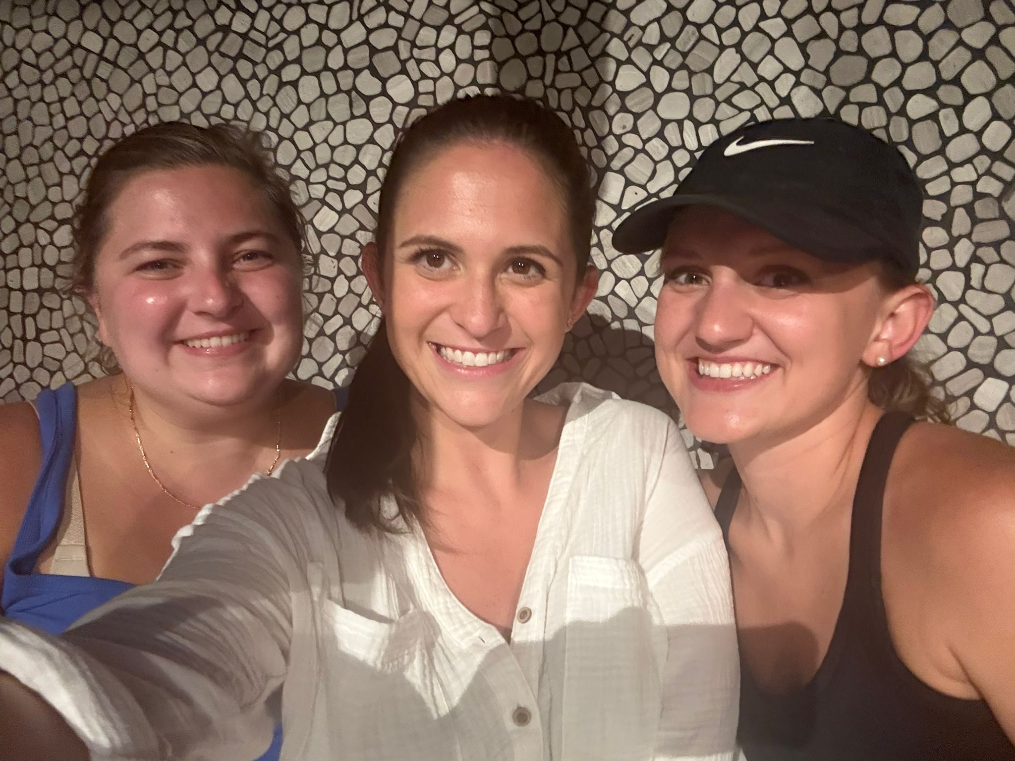 Three women smiling in a selfie against a stone-patterned wall. The tone is warm and joyful, with casual attire and relaxed expressions.