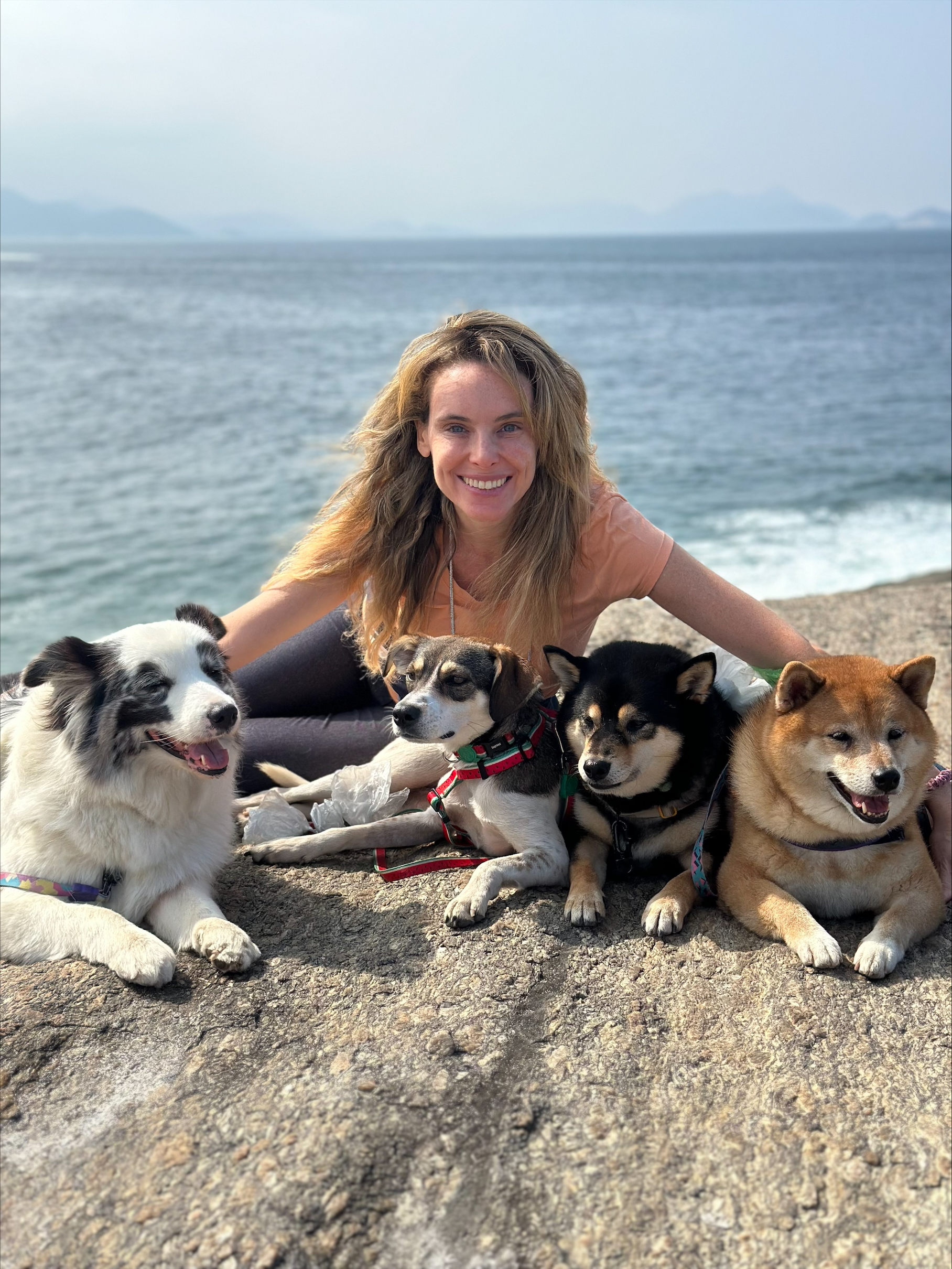 Luiza Goldemberg sits on a rocky shore with four dogs by the sea. The scene is bright and serene, conveying happiness and companionship.