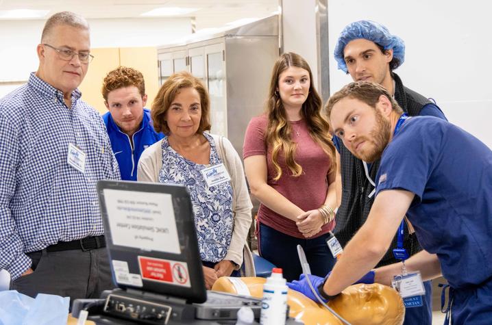 A group of five people attentively observes a medical simulation as an anesthesiology resident in scrubs demonstrates a procedure on a mannequin. The setting is clinical, with a focused and educational atmosphere.