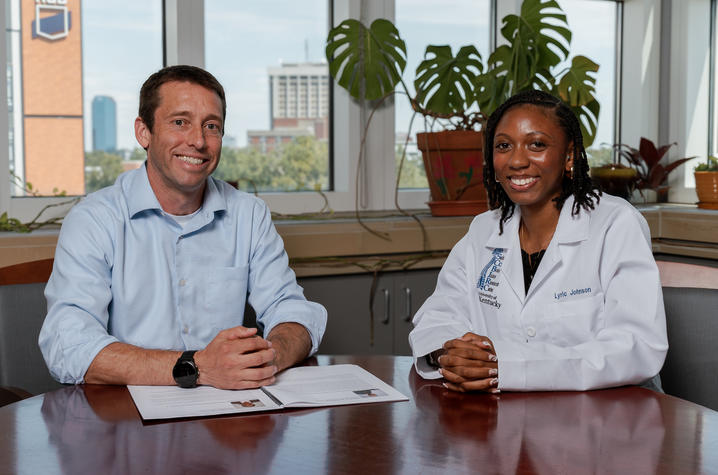 John Gensel and Lyric Johnson sit at a table in an office. John Gensel is in a light blue shirt, and Lyric Johnson, wearing her white lab coat, is sitting beside him. The two of them are smiling. A document lies on the table, and large potted plants are in the background, creating a warm professional atmosphere.