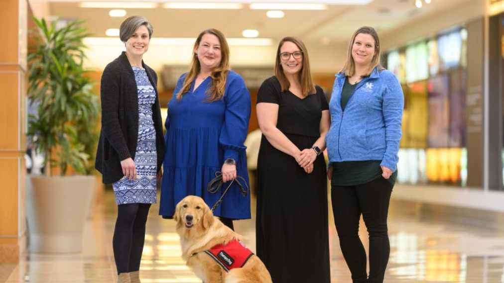 Four women stand together smiling in a bright hallway, with a golden retriever wearing a red therapy dog vest sitting at their feet.