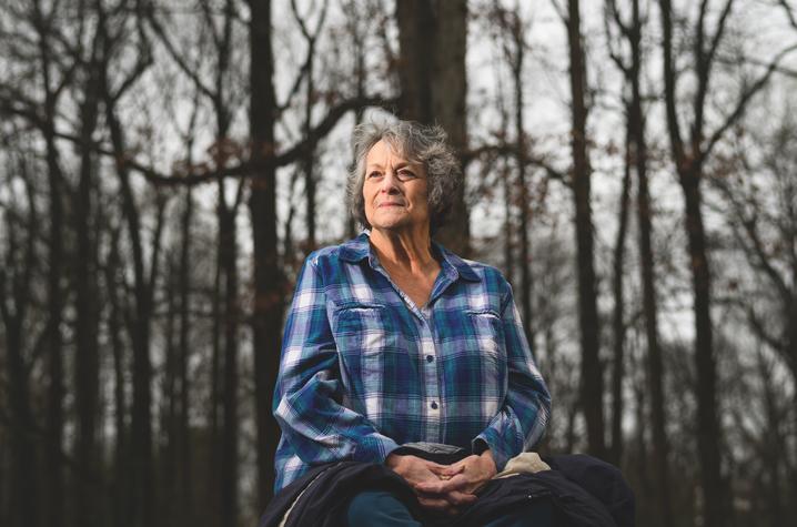 An elderly woman with short, grey hair, wearing a blue plaid shirt. She is sitting outdoors in front of a tree-line, with hands in her lap. She smiles softly into the light of the setting sun.