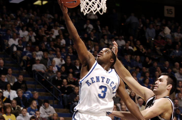 Brandon Stockton in a white Kentucky jersey leaps toward the basket for a layup, with an opposing player defending. The crowd watches intently.