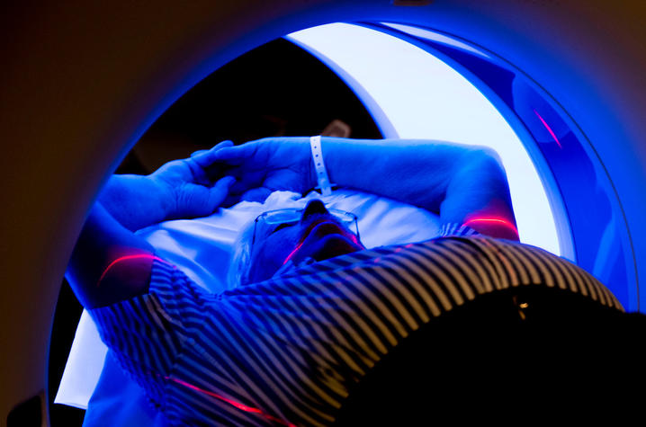 A man in a striped shirt, laying back and holding his arms over his head while receiving a CT scan.