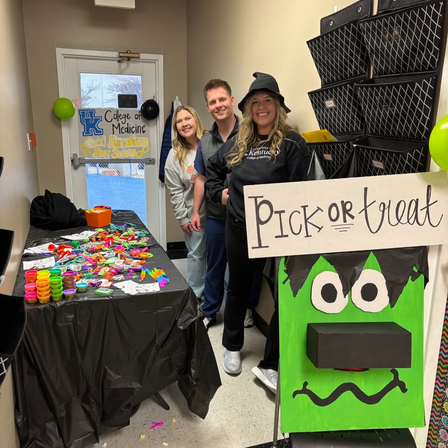 Three medical students dressed for Halloween pose next to a table full of toys and a sign that says "pick or treat" during a Halloween event