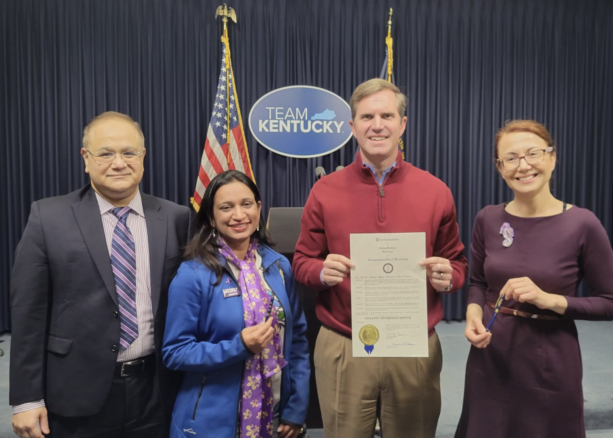 Three individuals from Neurology stand smiling with Governor Beshear in front of "Team Kentucky" signage and American flags. One holds a document, indicating an official or celebratory event.