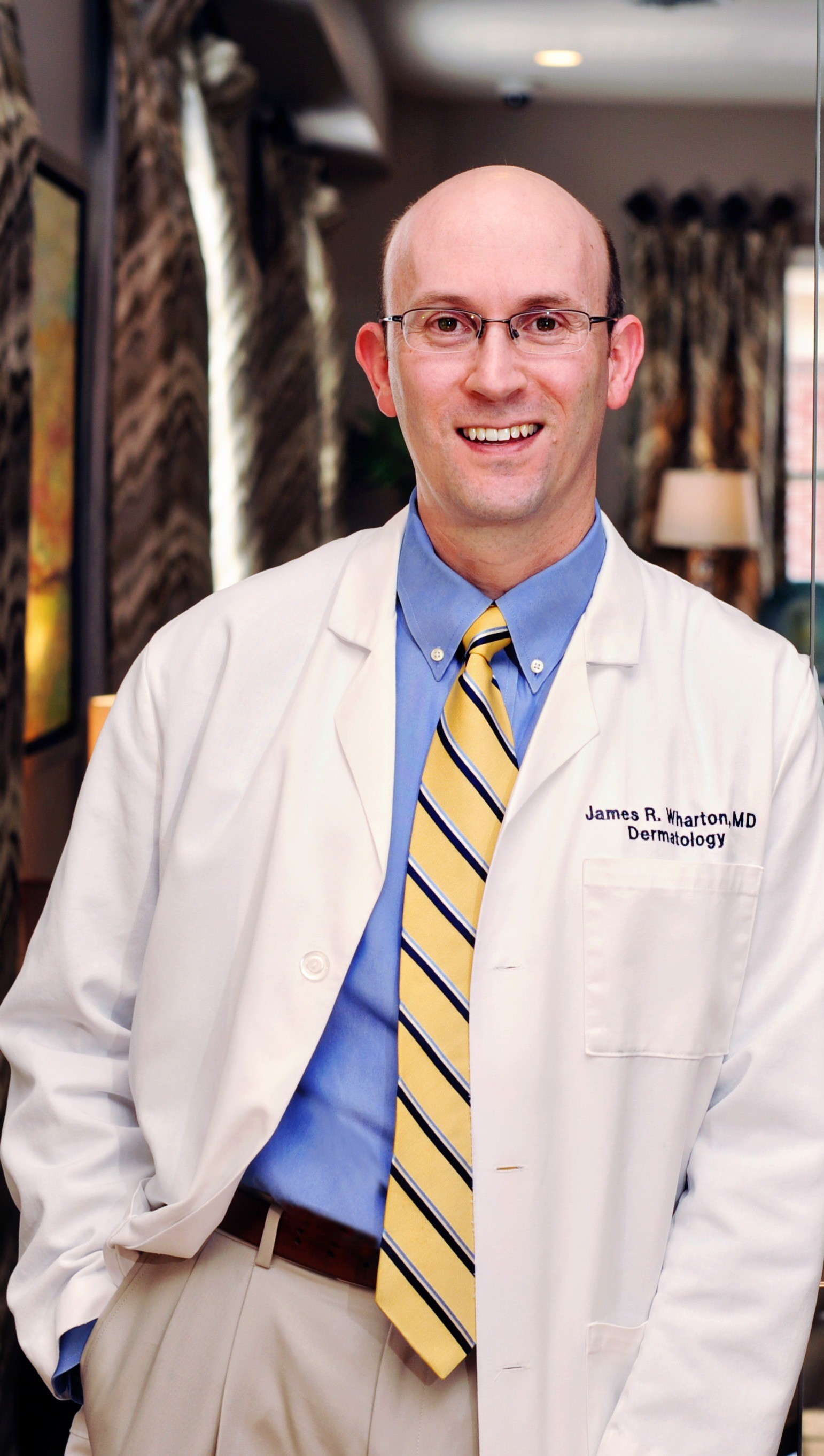 Dr. James Wharton posts for a headshot in a white coat, blue shirt, and striped, yellow tie
