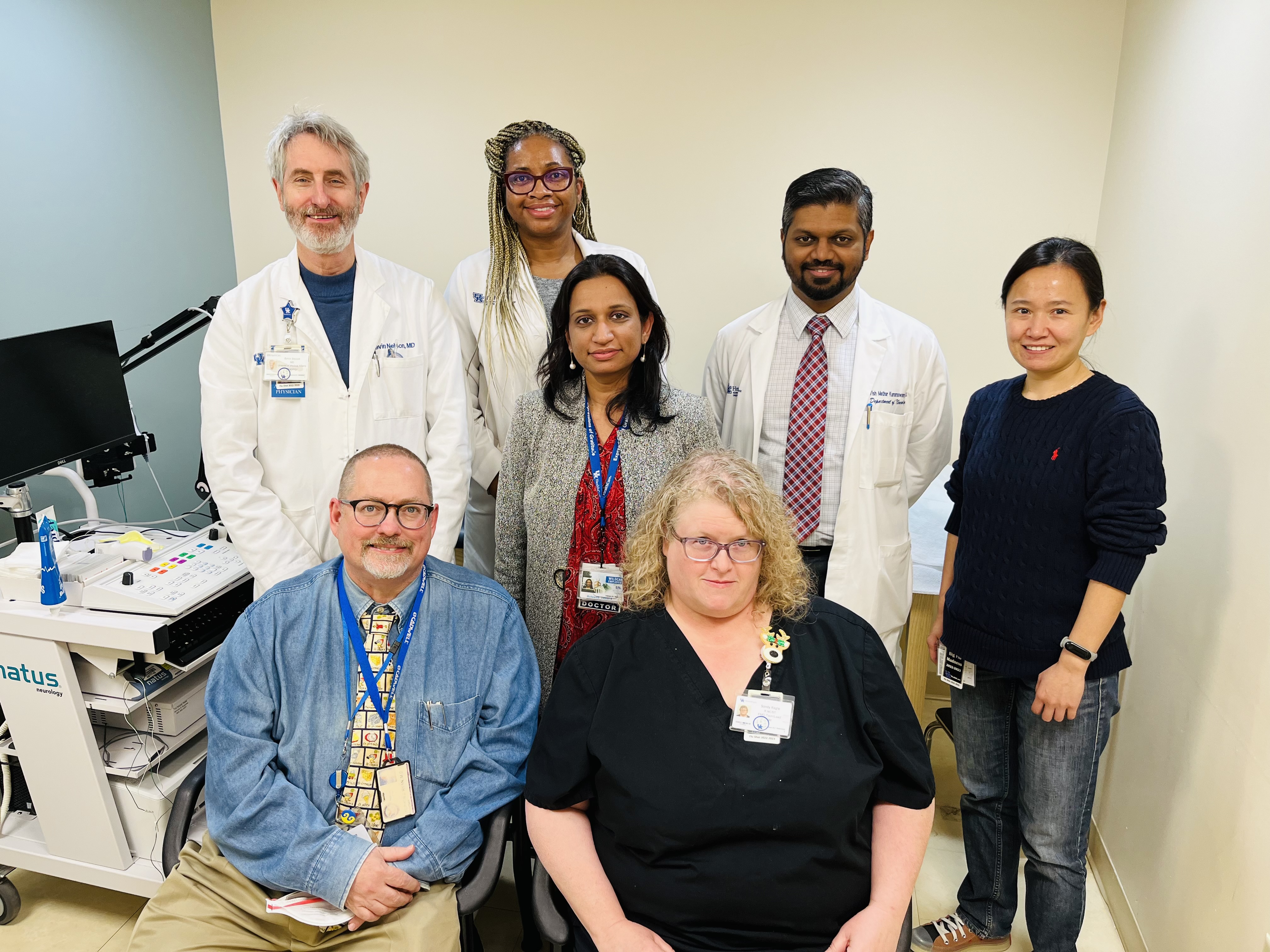 A group of seven medical professionals in a hospital setting, smiling. Three males and four females, wearing white coats and casual attire, suggesting teamwork.