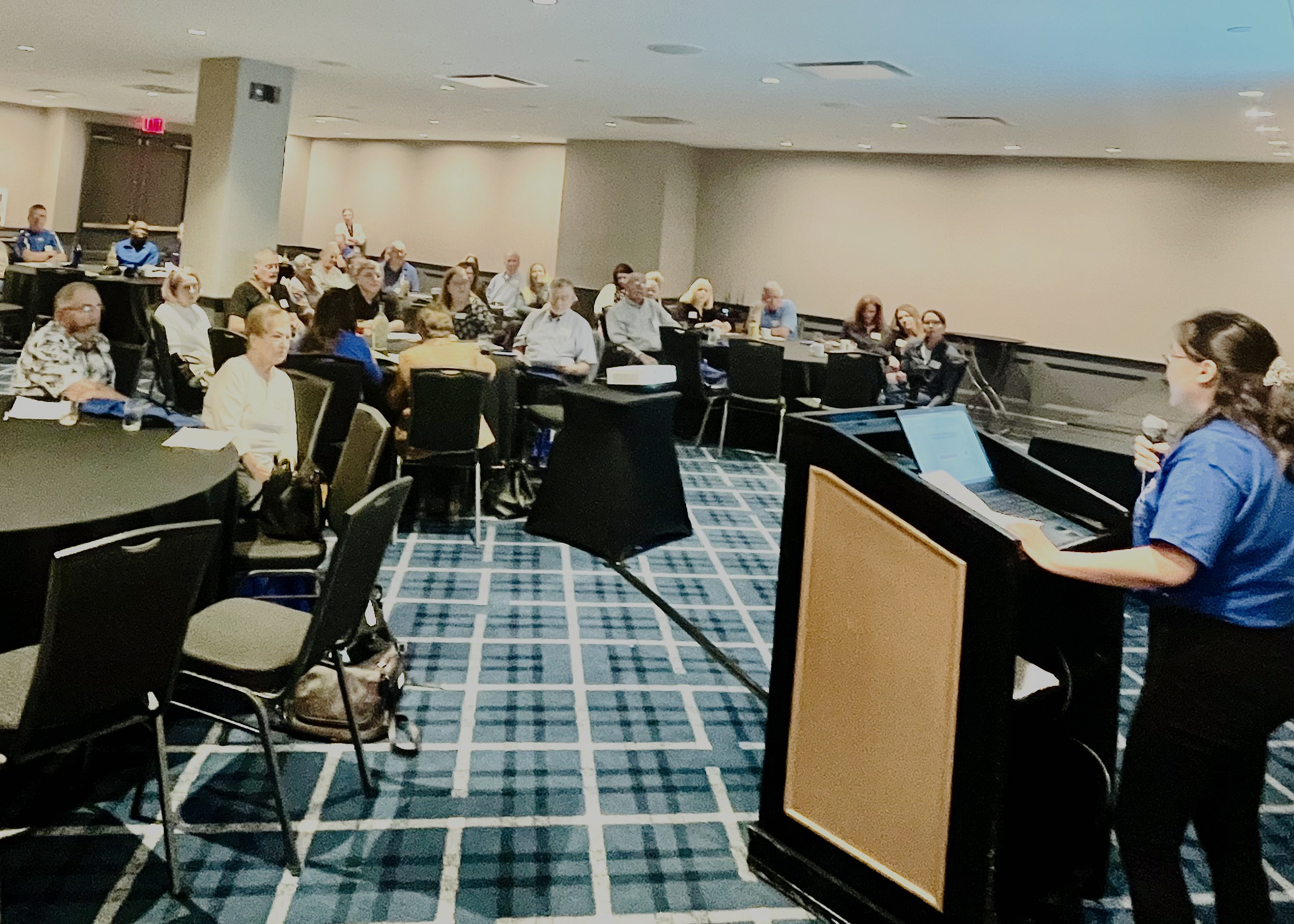 A woman in a blue shirt speaks at a podium to an attentive audience seated at round tables in a conference room. The atmosphere is focused and engaging.