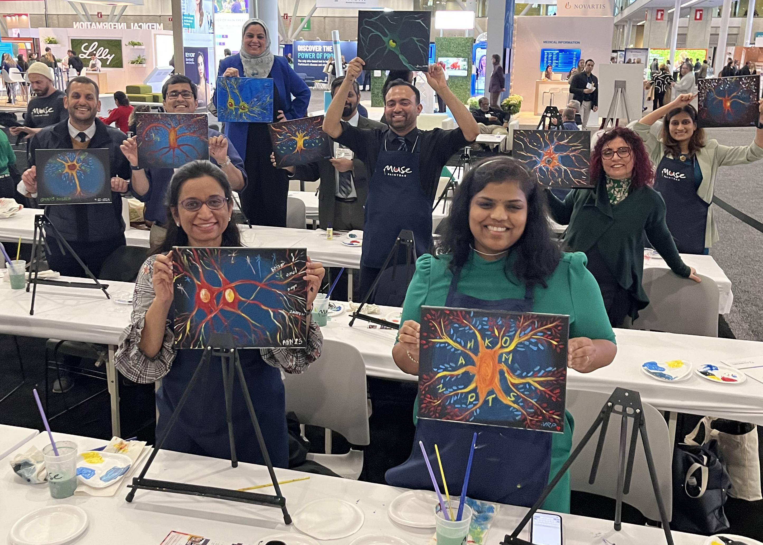A group of smiling people hold up their vibrant paintings of colorful neurons during a workshop. They are wearing aprons and standing in a brightly lit venue.