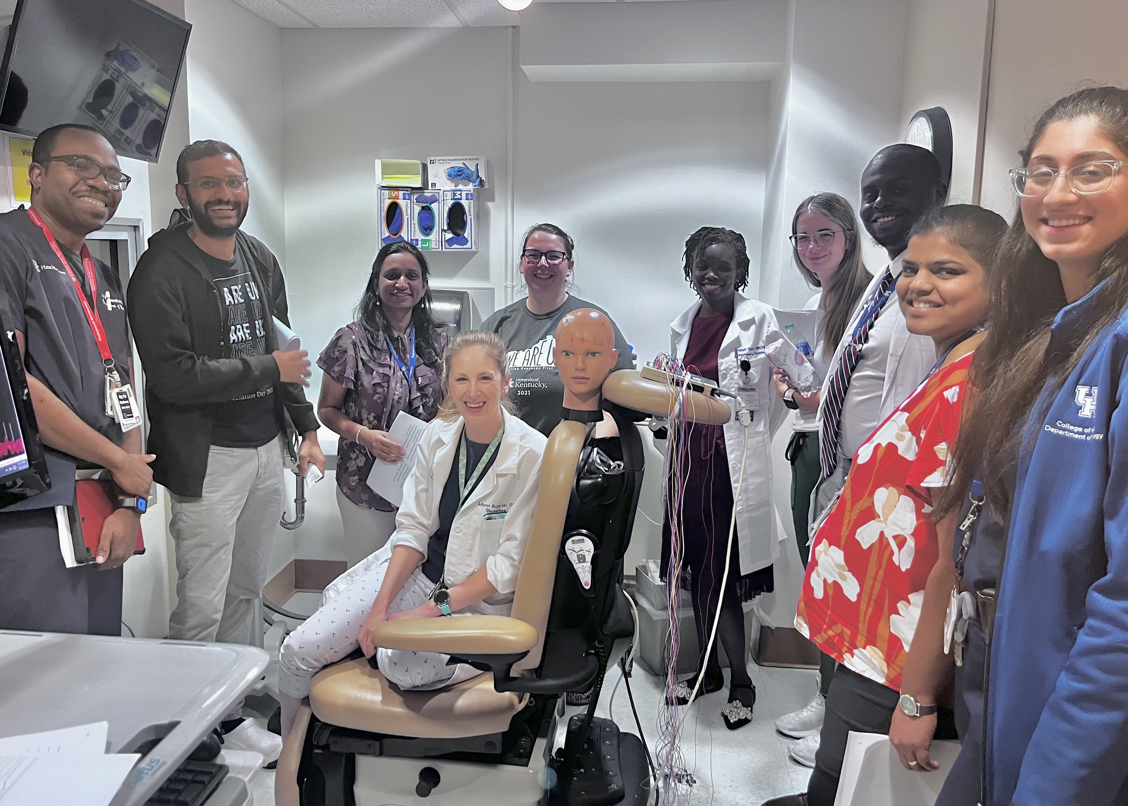 A diverse group of people in medical attire pose around a medical dummy in a clinical setting, conveying teamwork and learning in a supportive environment.