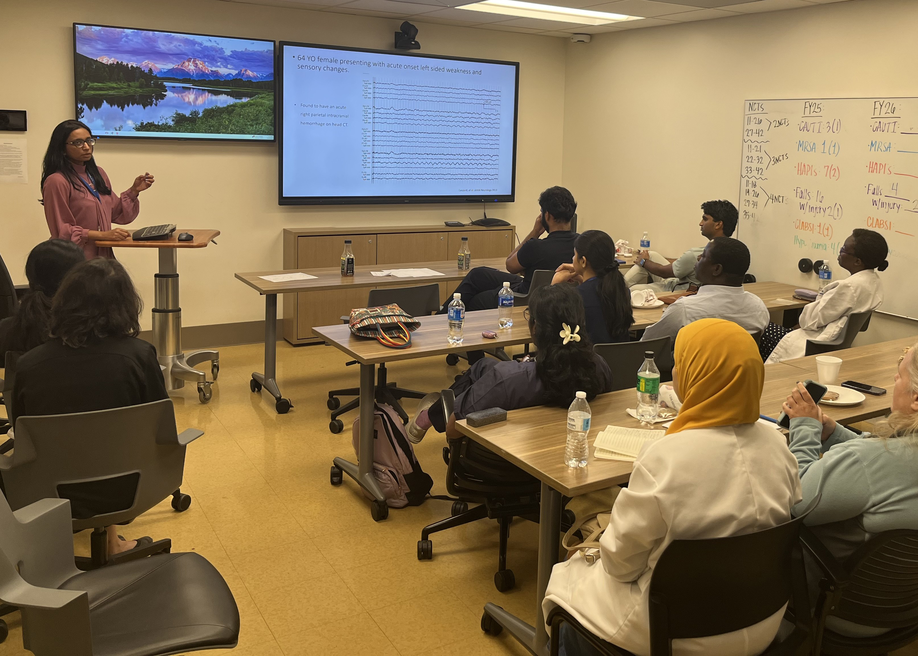 A woman presents in a classroom with attendees seated around tables. A screen shows presentation slides. The room is engaged and focused.