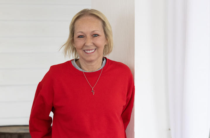 Vikki Lane, a blond woman wearing a red sweatshirt, leans against a wall and smiles. The image evokes positivity and happiness.