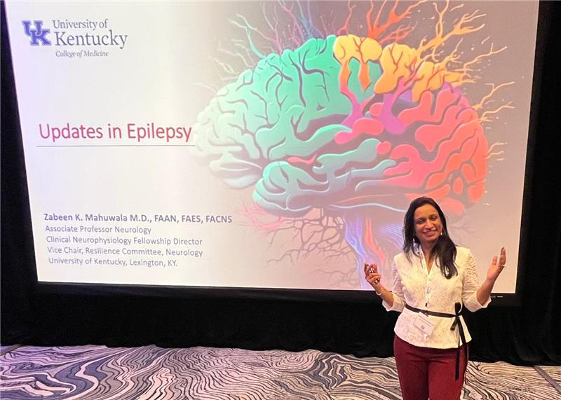 A woman in a white blouse stands smiling in front of a vibrant brain illustration on a presentation slide titled "Updates in Epilepsy" at a University of Kentucky event.