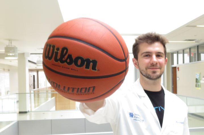 Bernardo Aguzzoli in a lab coat holds a Wilson basketball in an indoor hallway, smiling confidently. The setting suggests a fusion of science and sports.