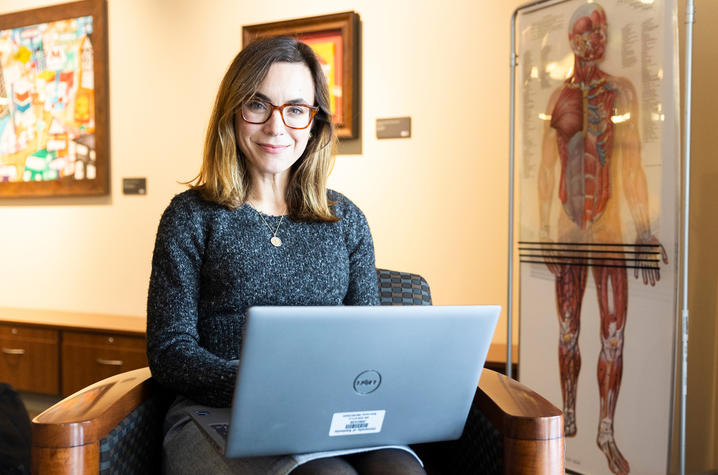 Sara Police sits in an office chair, smiling, and holding a laptop. The background features a colorful painting and an anatomical poster, creating a scholarly atmosphere.
