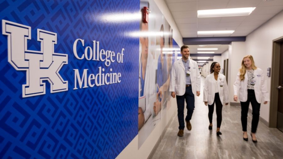 Three medical students wearing white coats walk down long hallway, UK College of Medicine logo to the left
