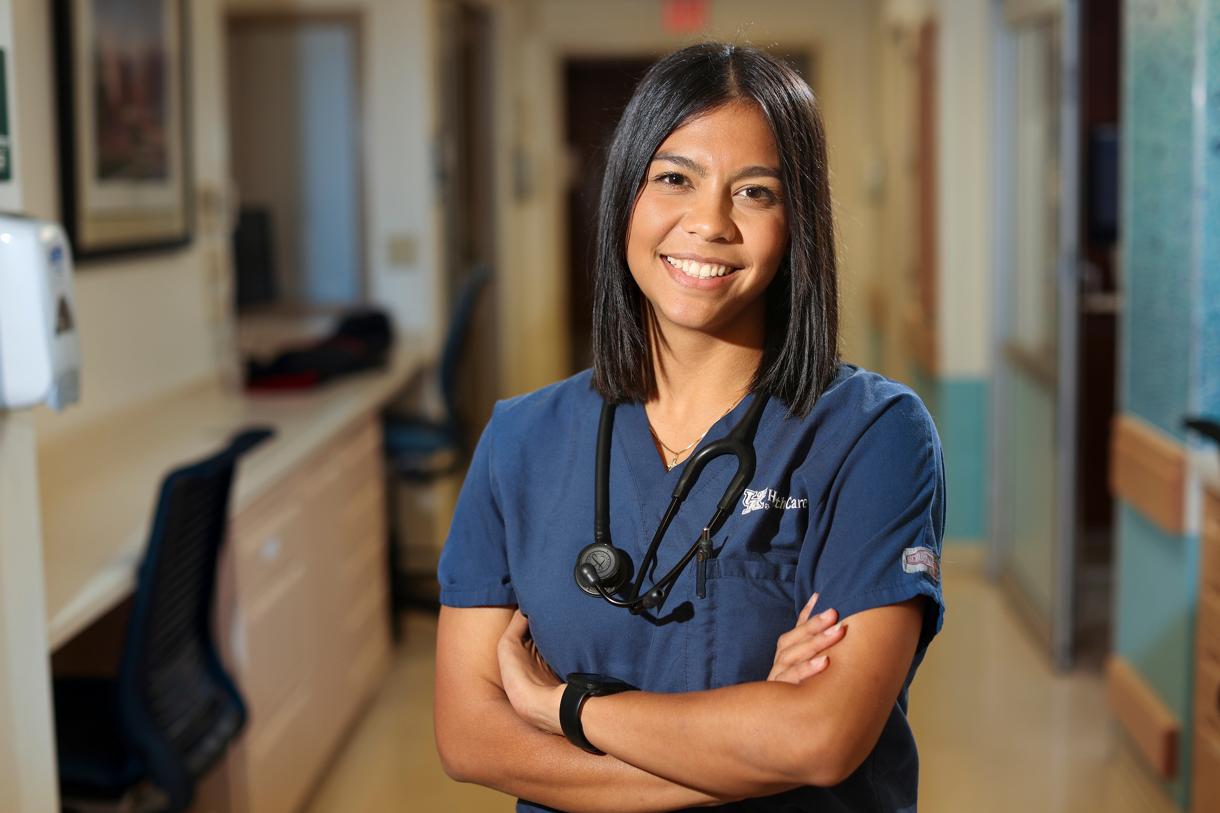 Smiling health care professional in navy scrubs with a stethoscope, standing confidently in a clinic hallway with arms crossed.