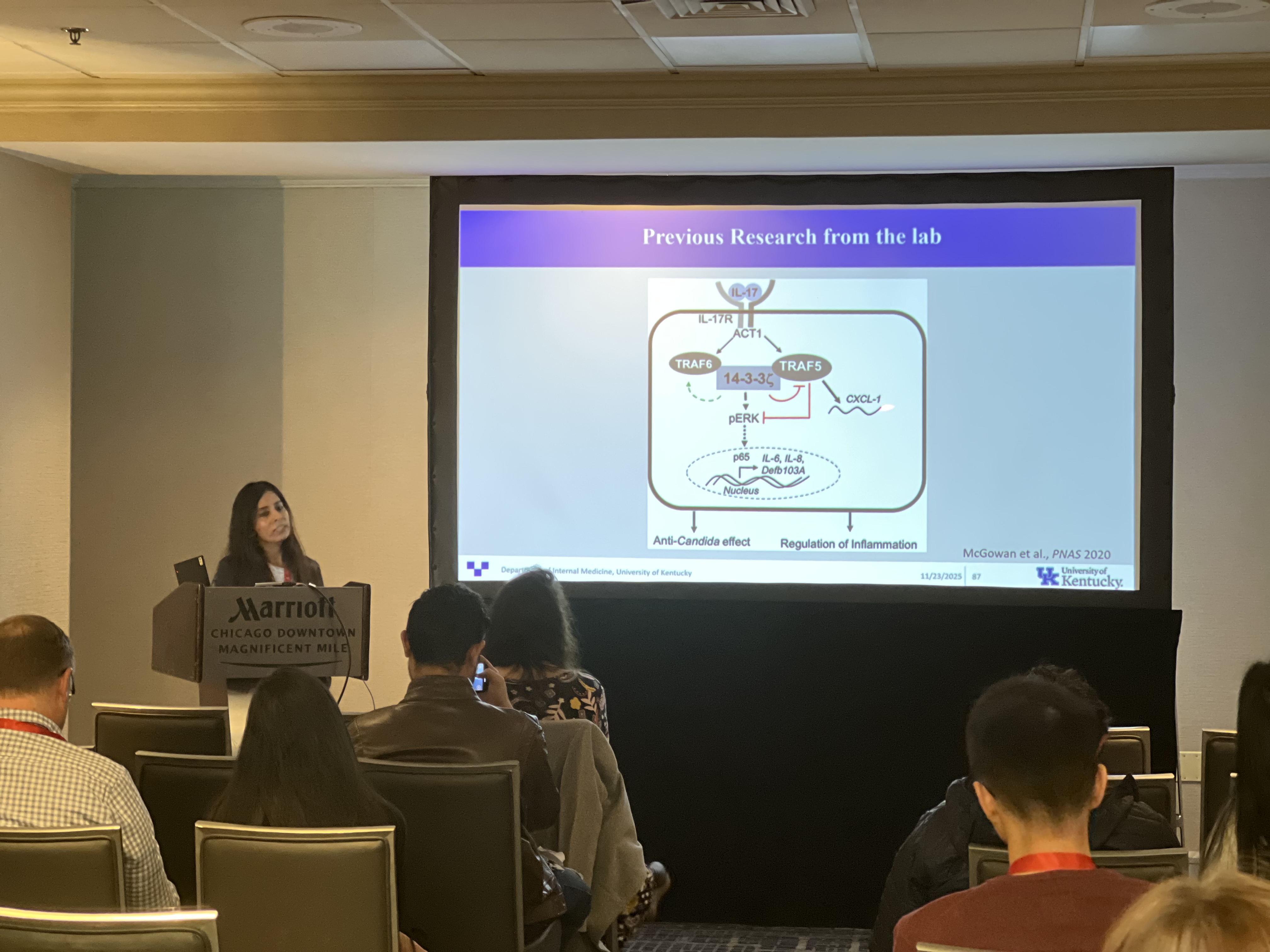 A woman presents in front of a group, standing next to a large projecter