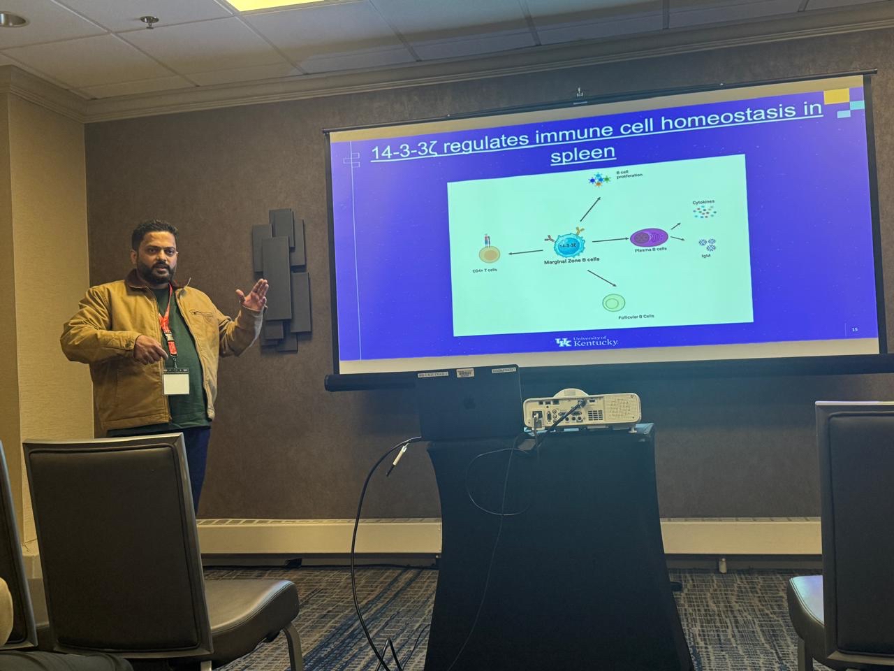 A man presents in front of a group, standing next to a large projector