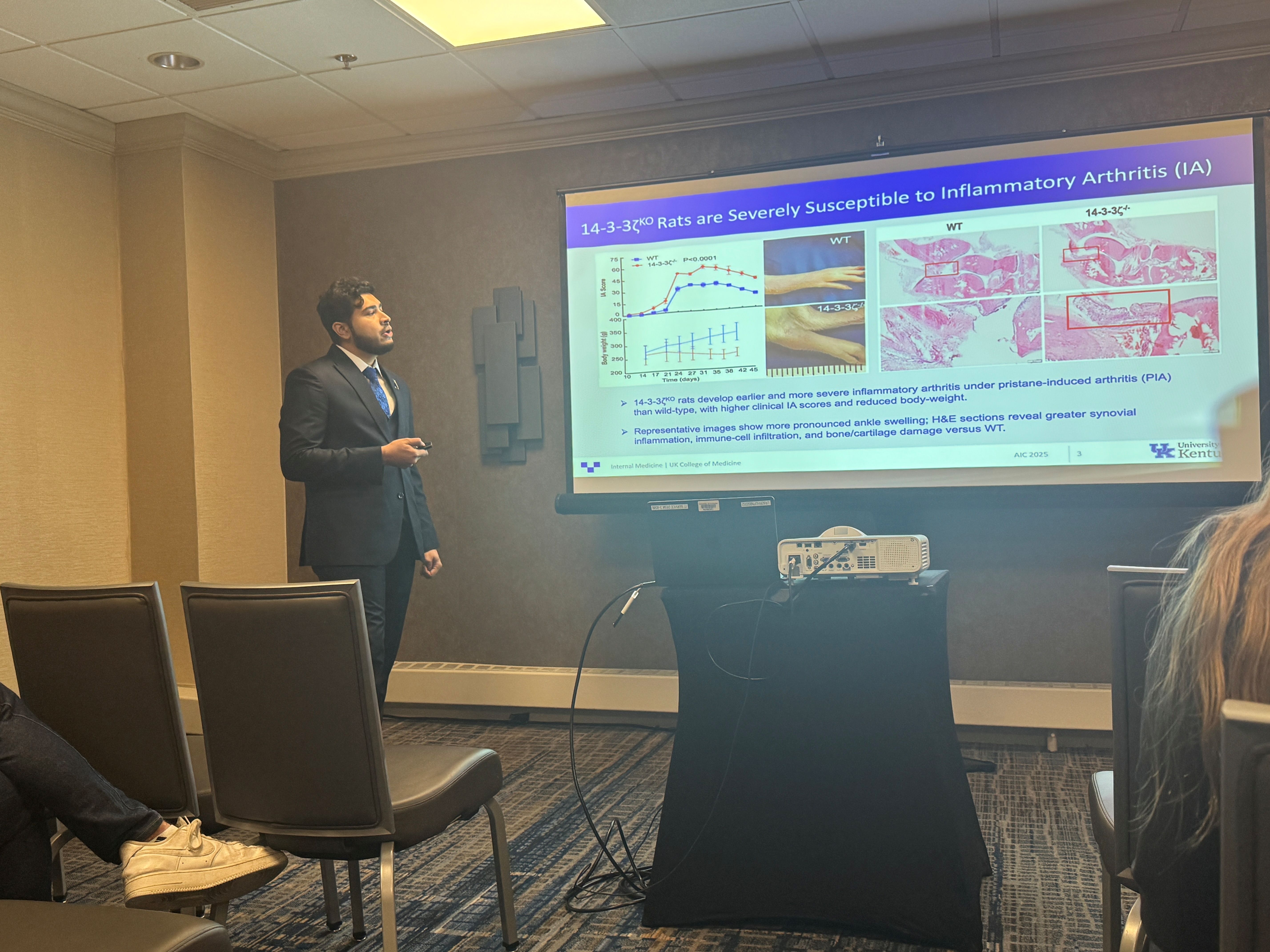 A man presents in front of a group, standing next to a large projector