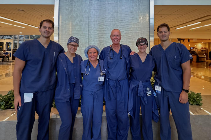 A smiling group of physicians stand in blue scrubs in front of a water wall in the UK hospital. The photo conveys positivity and community.