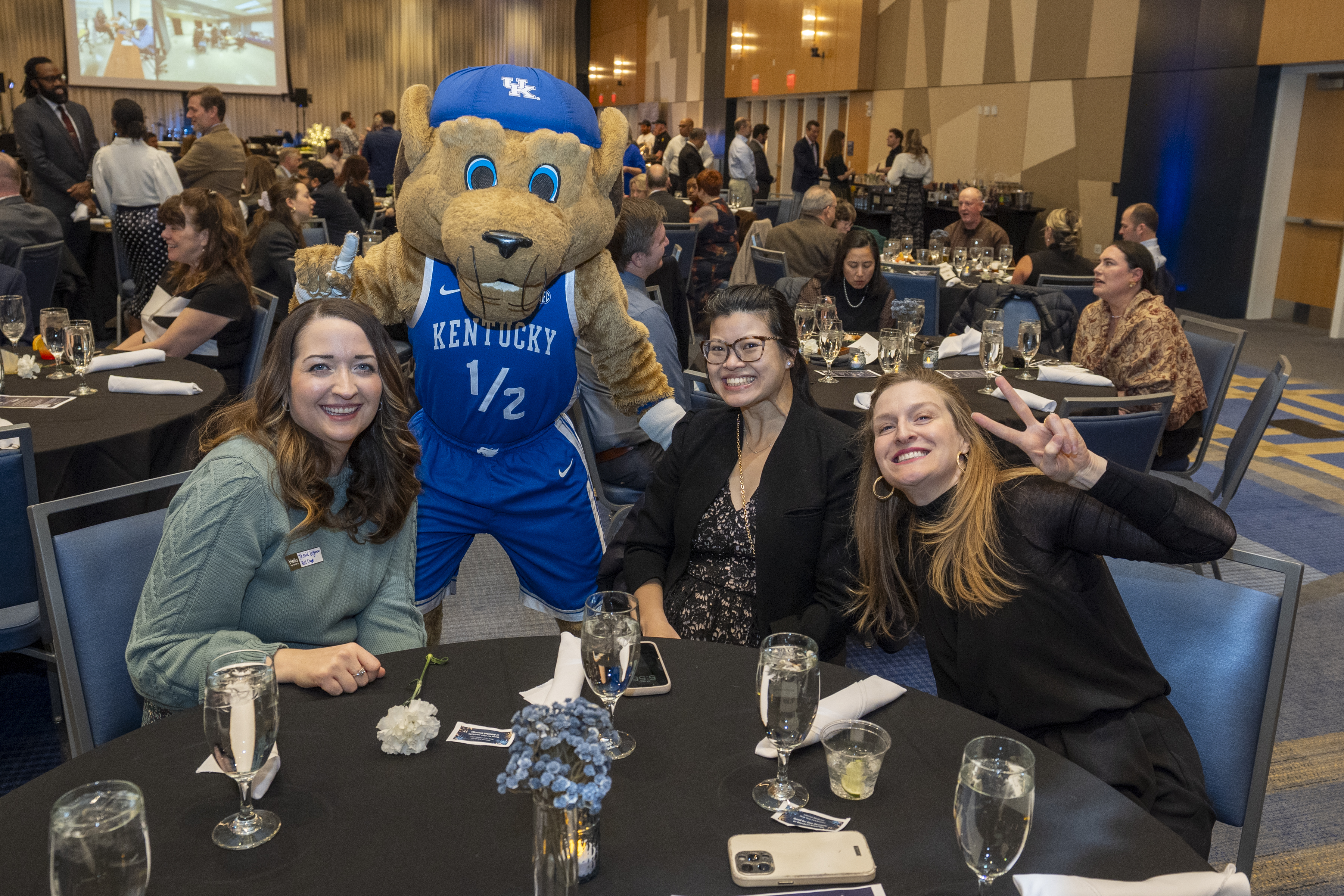 Three women sit at a table, posing with Scratch the wildcat. One woman throws up a peace sign