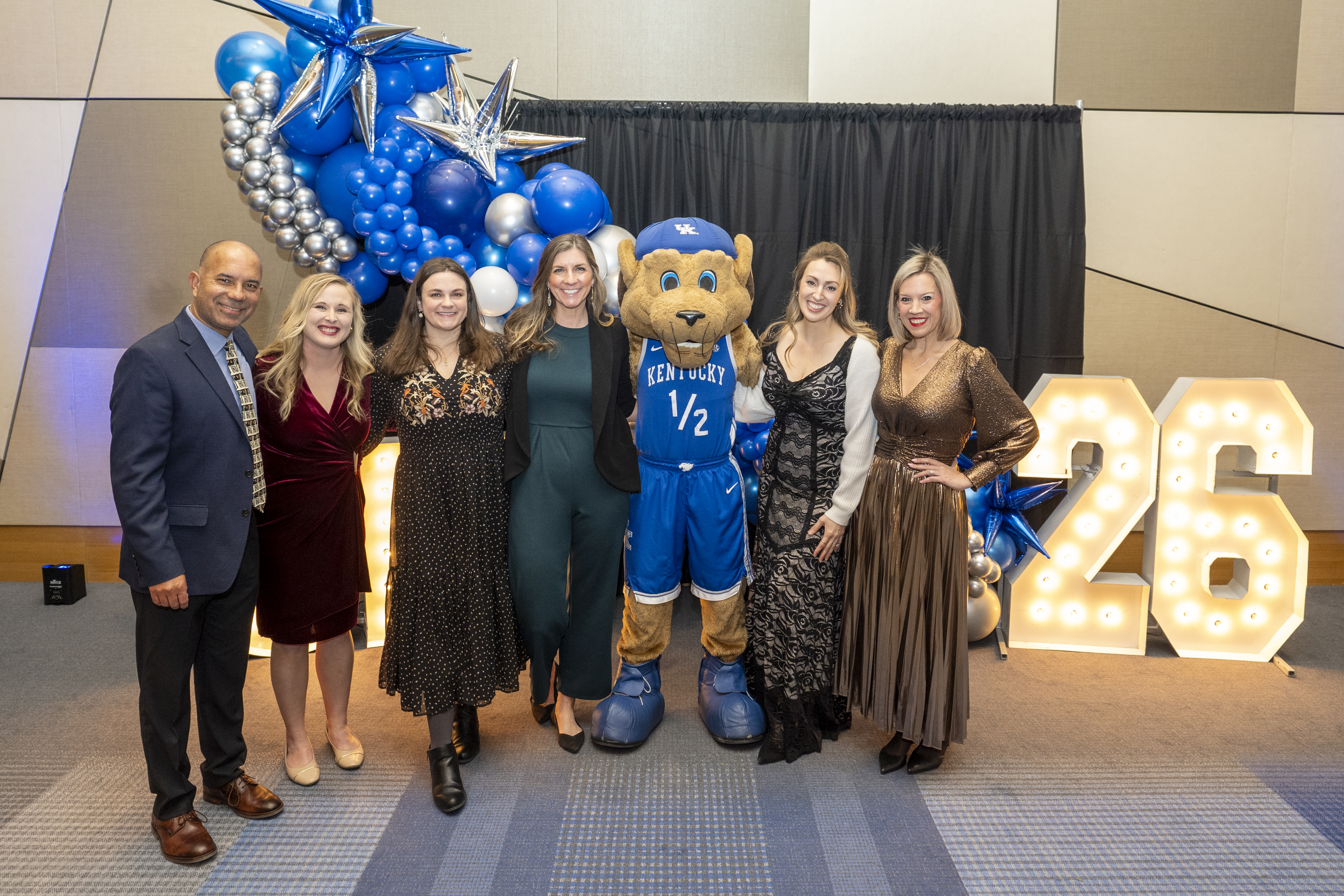 A group of people stand in formal attire with Scratch the mascot. They are next to large marquee letters that say 26