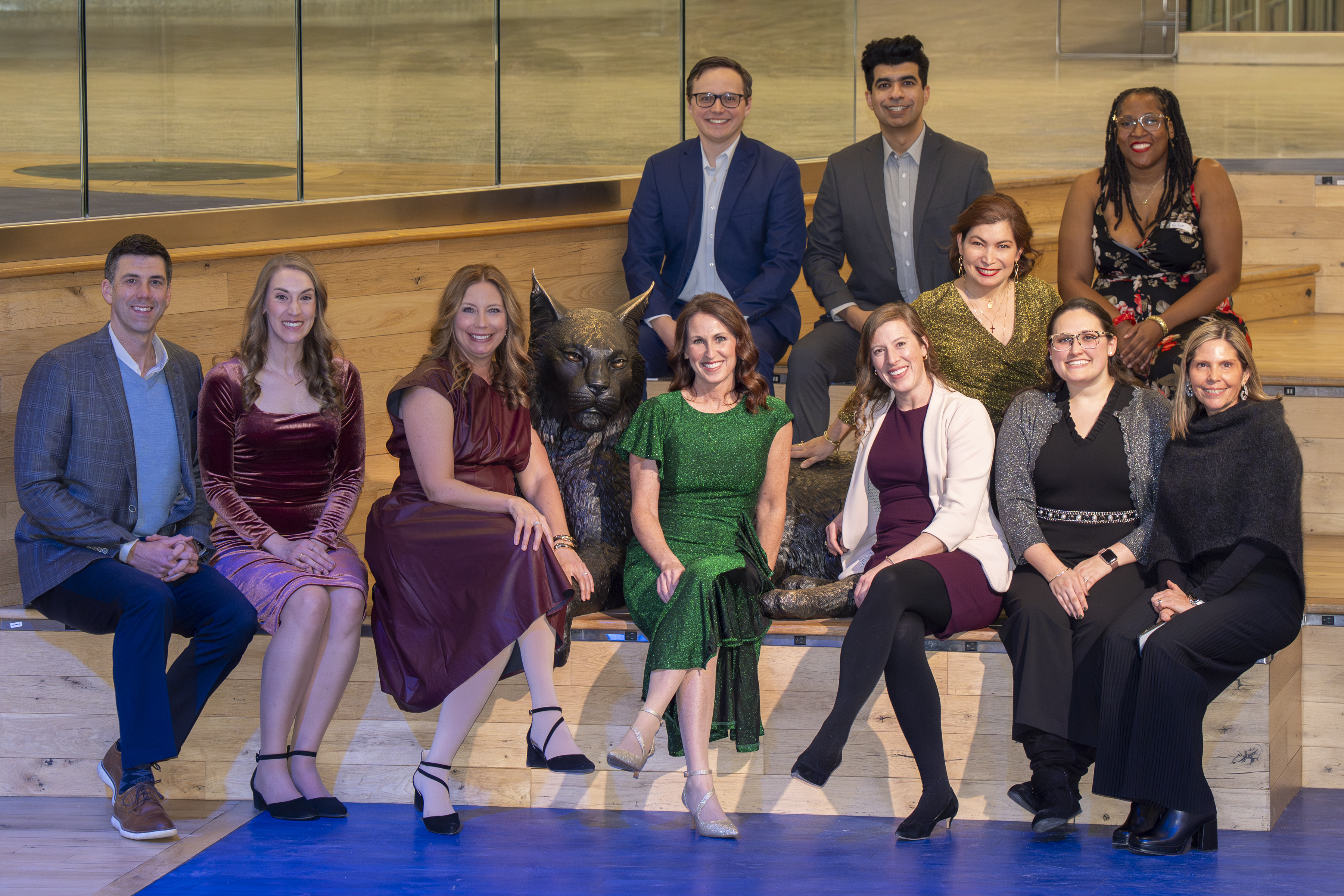 A group of folks in formal attire smile on a large, wooden stair case around a metal statue of a wildcat