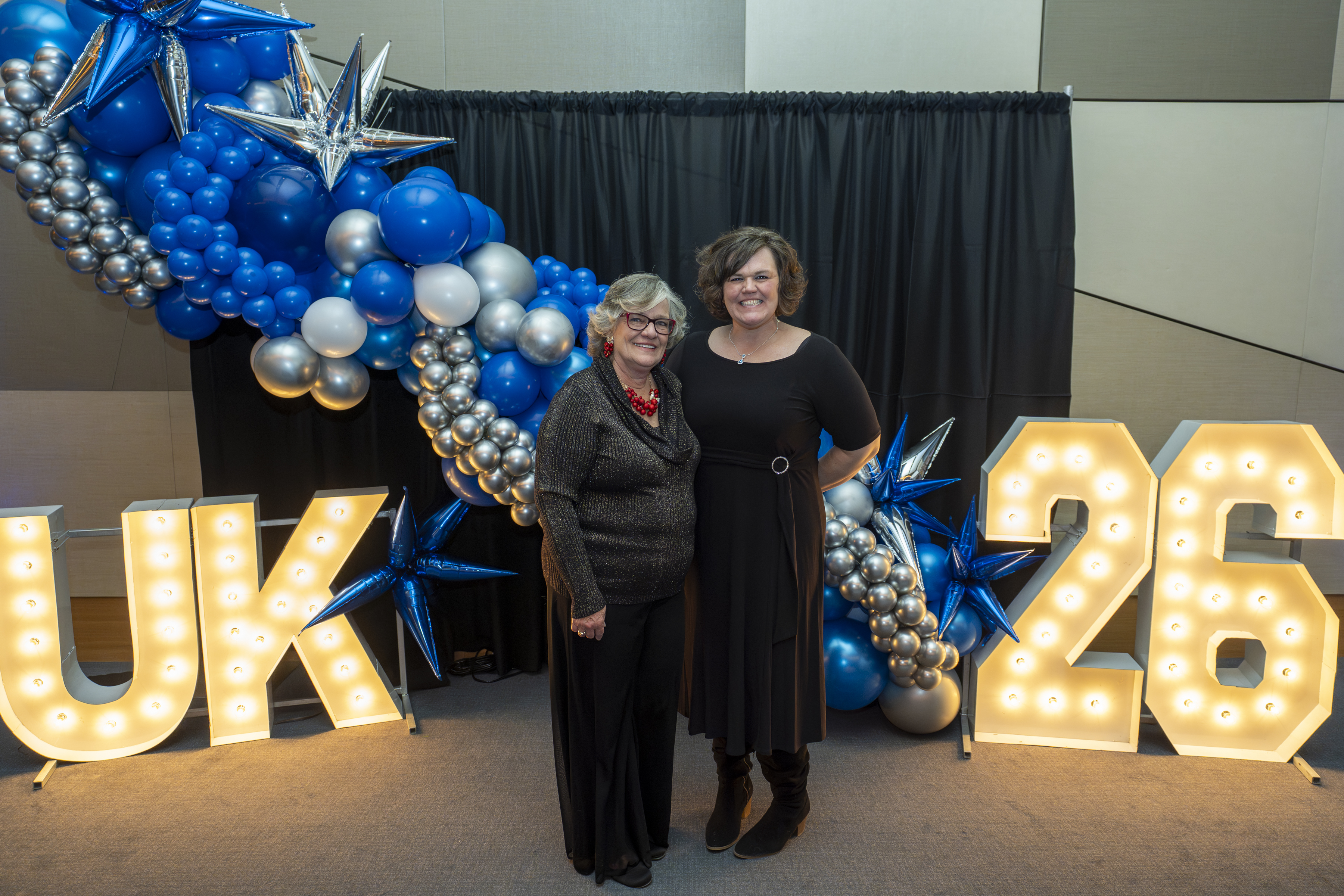 Two women stand together between large marquee letters spelling UK and 26