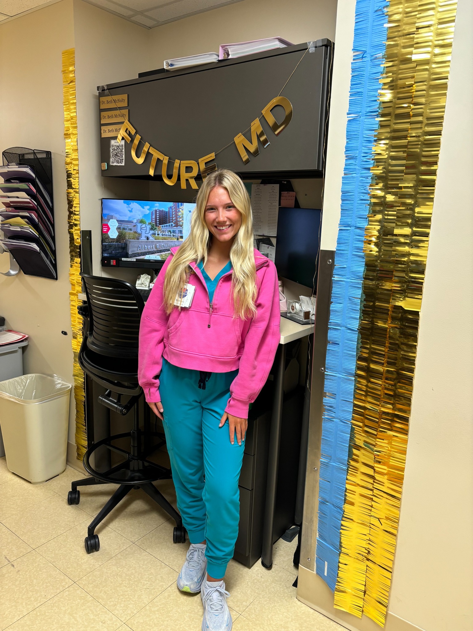 A woman in a pink hoodie and teal scrub pants smiles in an office decorated with "Future MD" banner. The room has gold and blue streamers, creating a celebratory tone.