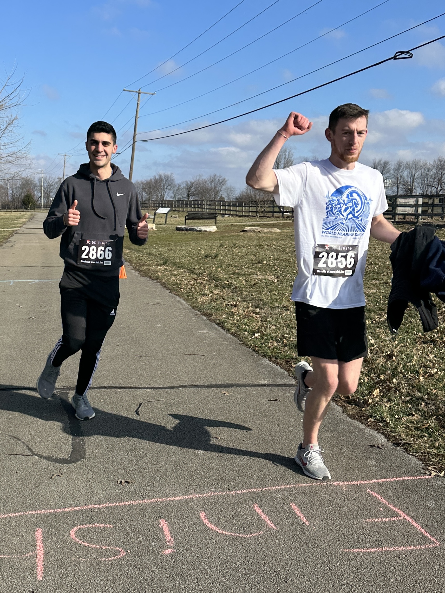 Two men finish a race on a sunny day. The one on the left smiles and gives a thumbs up, while the other raises his arm in triumph. "Finish" is marked on the path.