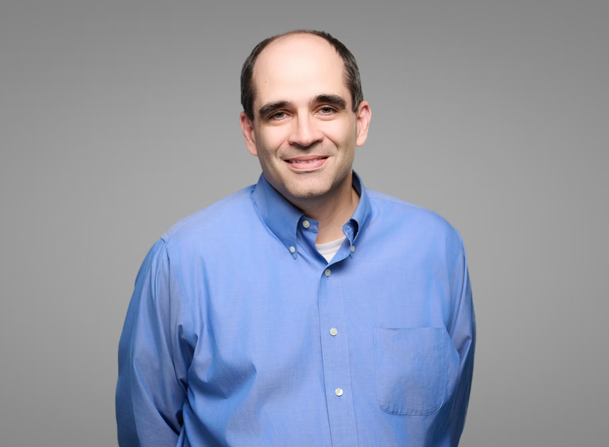 Headshot of Dr. Penaskovic, wearing a light blue button-up shirt in front of a gray background, smiling gently. 