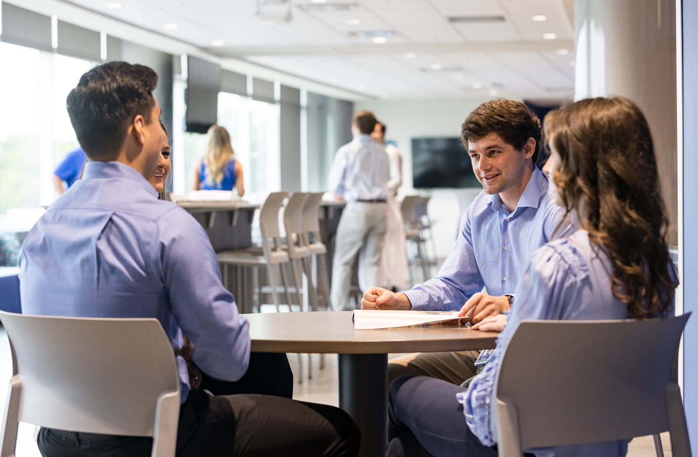 Four people in a modern office lounge sit around a table in discussion, smiling. Others stand and chat in the background.
