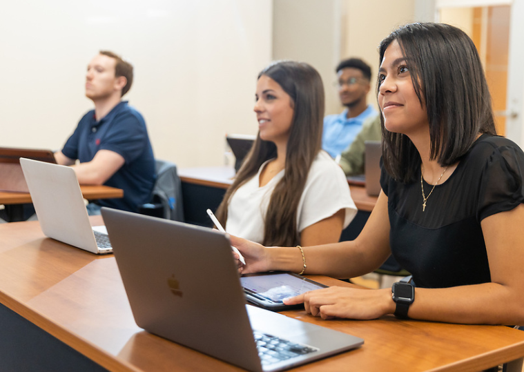 Students in a classroom, attentively listening and taking notes on laptops. 