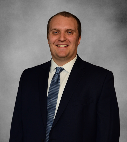 Dr. Andrew Arnold poses for professional headshot wearing a black suit and blue tie in front of a gray background.