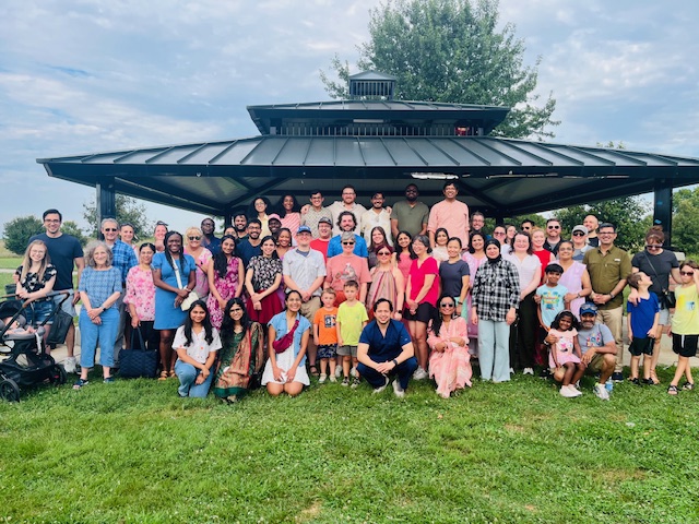 A group of Neurology members and their families standing under a park shelter on a sunny day. The photo conveys camaraderie and joy.