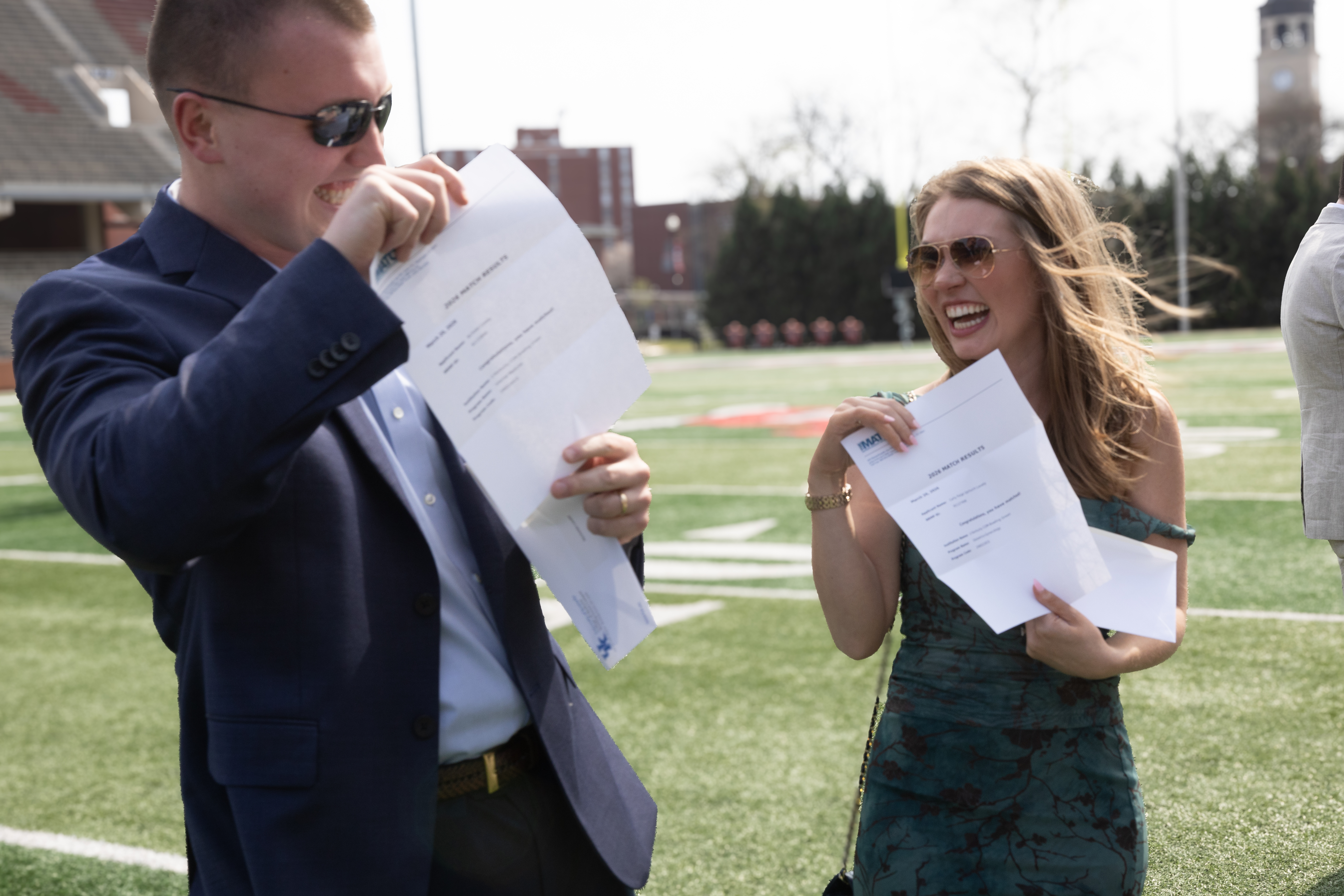 Nick and Carly Lovely joyfully hold their Match letters on a sunny day in a stadium. The setting exudes excitement and accomplishment.